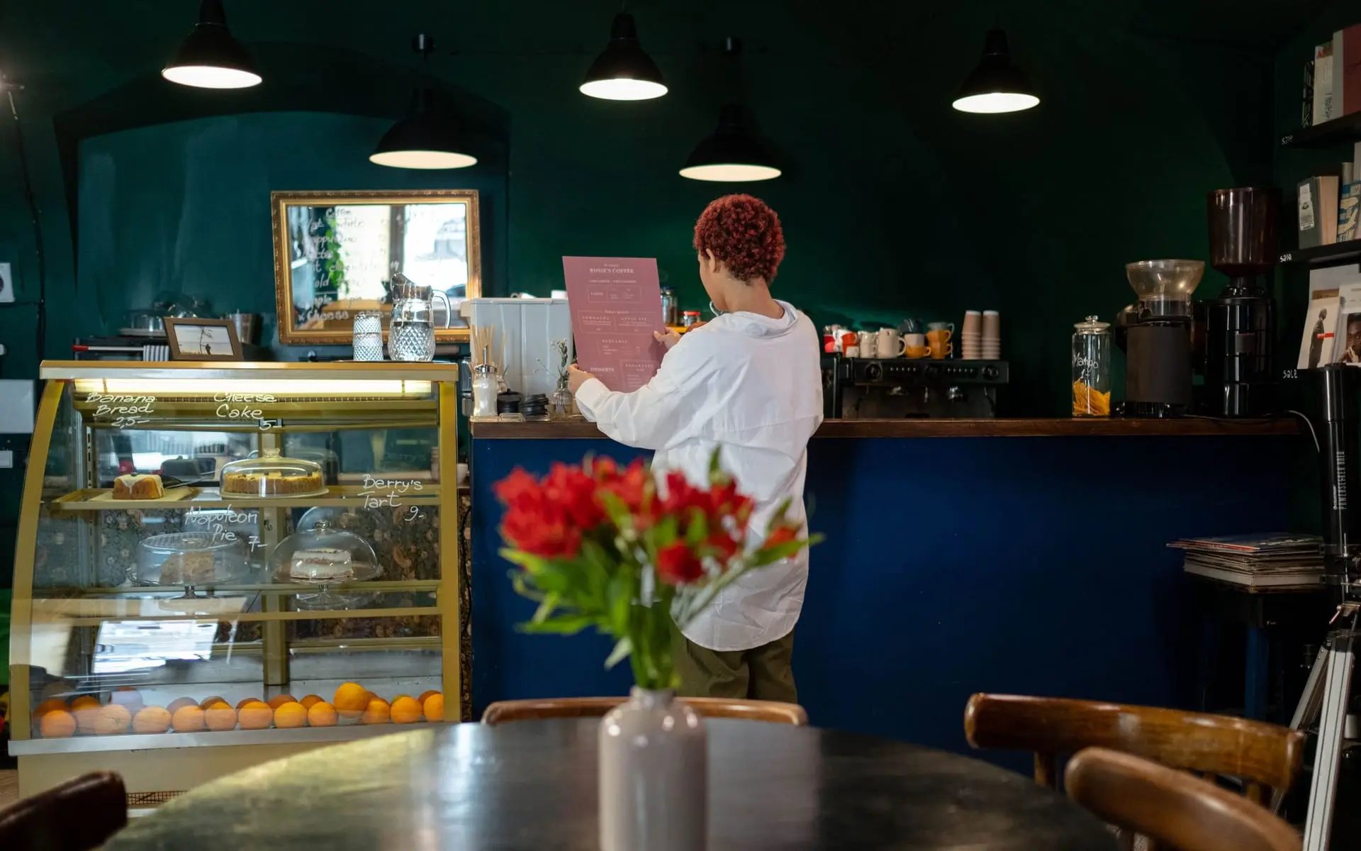 Cafetería de barrio con vitrina de postres, barra azul y una empleada atendiendo al fondo