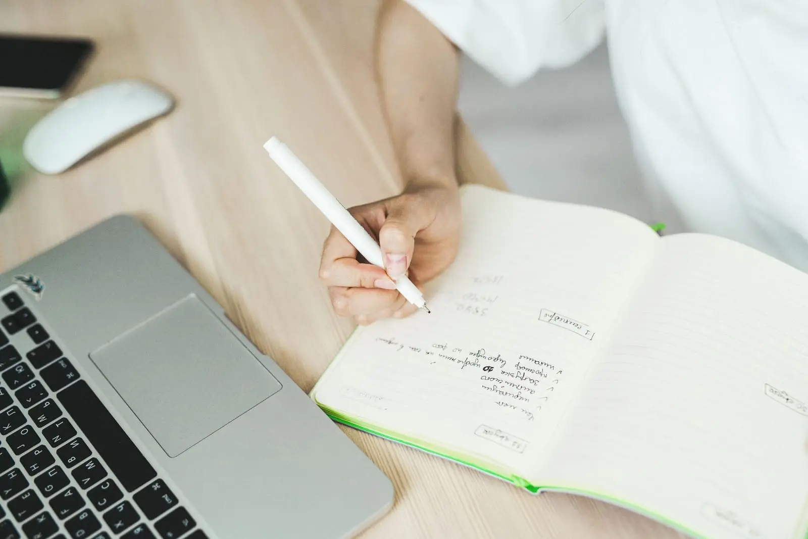 Mano con bolígrafo blanco anota una lista a mano en una libreta abierta junto a un portátil