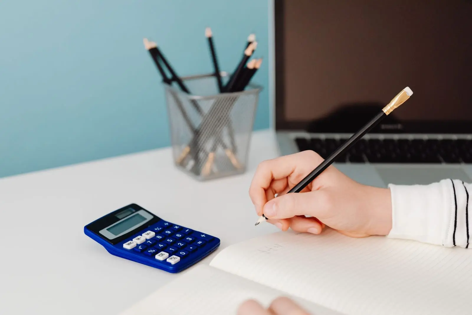 Mano que escribe con lápiz en un cuaderno junto a una calculadora azul sobre un escritorio blanco