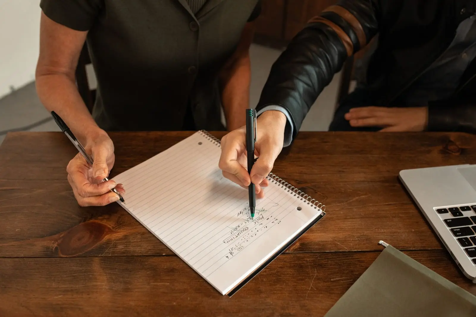 Dos personas anotan a la vez en una libreta de espiral sobre una mesa de madera