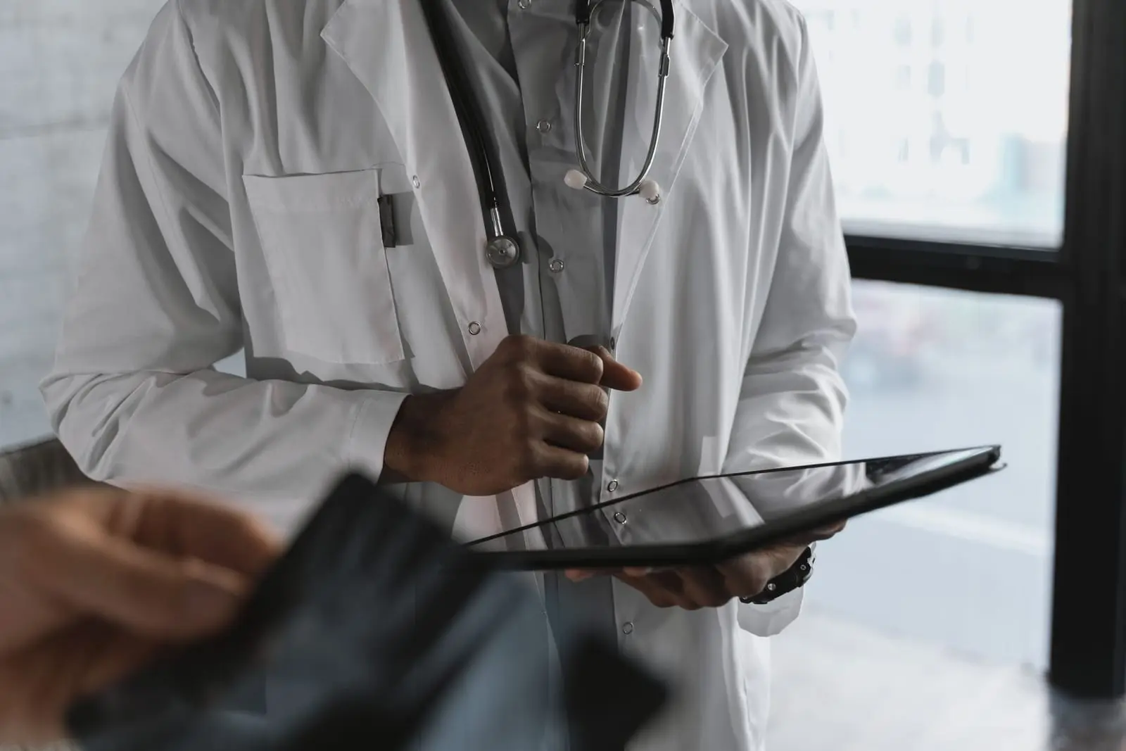 Médico con bata blanca y fonendoscopio consulta una tableta, sin mostrar el rostro