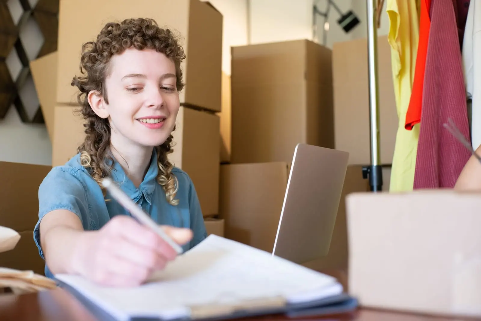 Emprendedora sonriente anota en una carpeta con cajas de envío y un portátil detrás